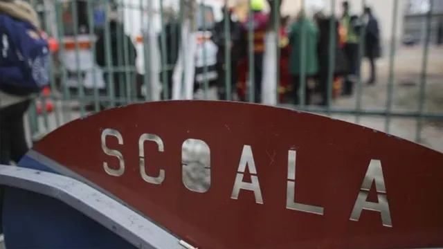 Un semn roșu cu textul metalic SCOALA este plasat în prim-plan, în fața unui gard metalic și a unor persoane neclare în fundal.