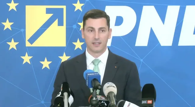 Un bărbat cu cămașă albă, cravată deschisă și sacou închis stă în fața unui fundal albastru cu stele galbene și litere mari albe.