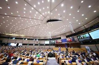 O sală de conferințe mare cu mulți oameni așezați la mese lungi dotate cu monitoare și un tavan înalt cu numeroase lumini punctiforme.