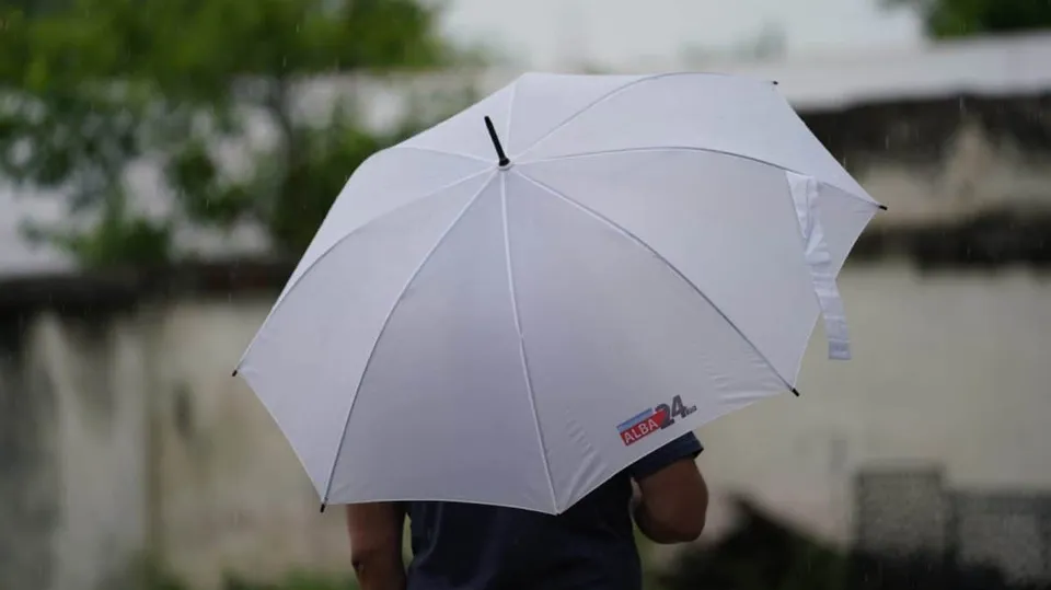 O persoană cu o cămașă închisă poartă un umbrelă albă sub ploaie în fața unui fundal cu vegetație și un perete alb.