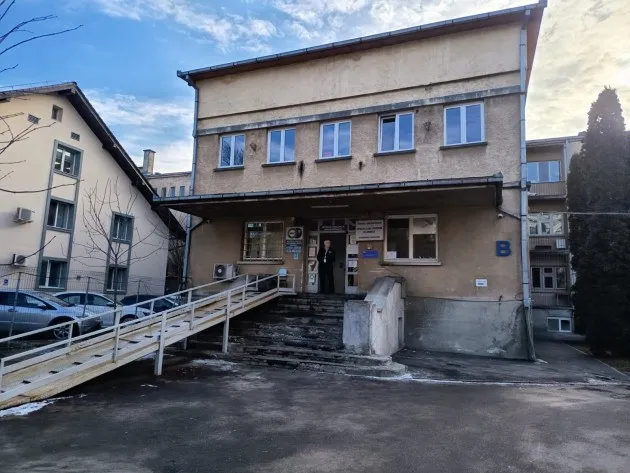 O clădire cu două etaje prezintă o rampă de acces din lemn, scări de piatră și ferestre cu rame albastre sub un cer înnorat.