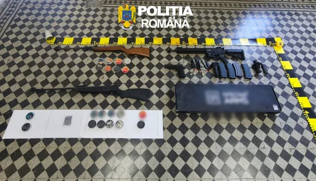 Arme de foc, muniție și un obiect dreptunghiular negru sunt dispuse pe un podea cu model de pătrate negre și albe, delimitată de o bandă galbenă.