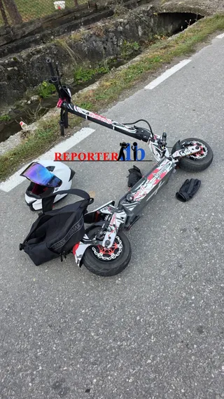 O trotinetă electrică cu modele colorate, o cască albă cu vizieră și o geantă neagră sunt așezate pe un drum asfaltat lângă un șanț.
