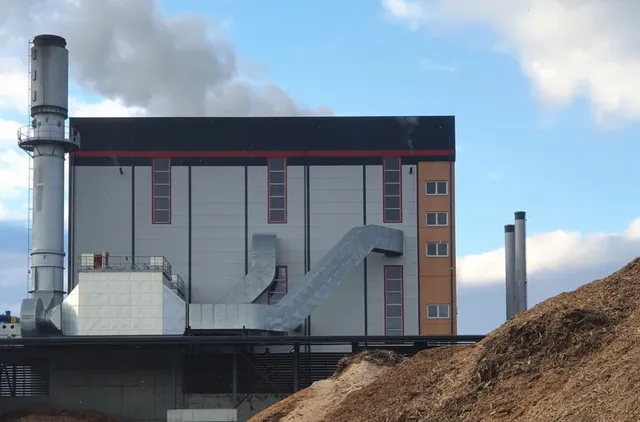 O clădire industrială gri cu detalii roșii și portocalii este situată lângă un coș înalt sub un cer cu nori.