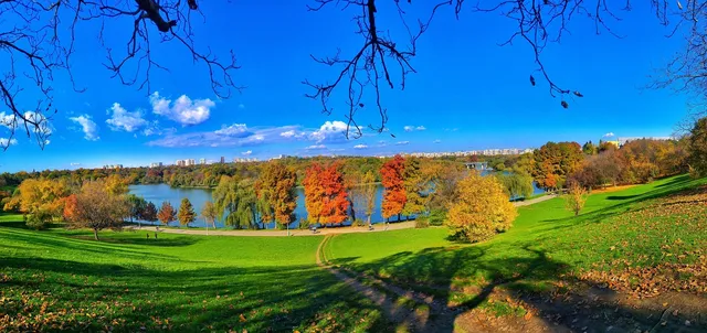 O vedere panoramică asupra unui parc cu iarbă verde, copaci cu frunziș colorat și un lac sub un cer albastru cu nori albi.