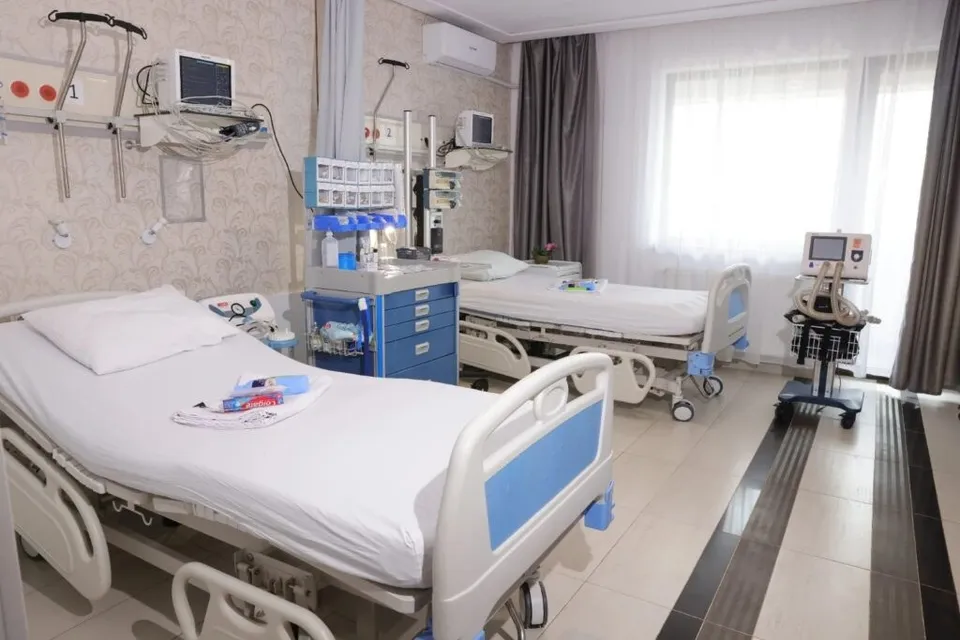 Două paturi de spital cu lenjerie albă sunt amplasate într-o cameră cu echipamente medicale și ferestre acoperite de draperii.