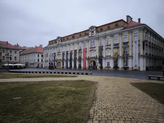 Un clădire mare, galbenă și gri cu ferestre multiple se află în spatele unui teren de iarbă și a unui drum pavat.