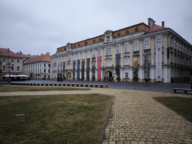 Un clădire mare, galbenă și gri cu ferestre multiple se află în spatele unui teren de iarbă și a unui drum pavat.