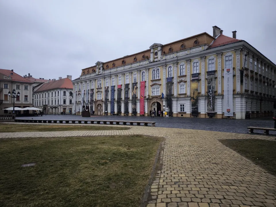 Un clădire mare, galbenă și gri cu ferestre multiple se află în spatele unui teren de iarbă și a unui drum pavat.