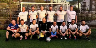 Un grup de oameni în echipamente sportive albe și negre pozează împreună pe un teren de fotbal verde în fața unei porți.