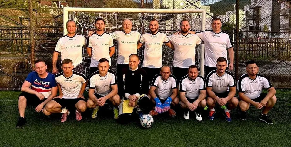 Un grup de oameni în echipamente sportive albe și negre pozează împreună pe un teren de fotbal verde în fața unei porți.