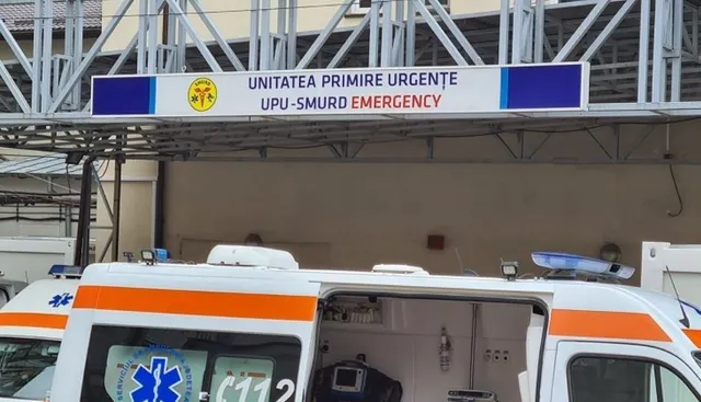 Imaginea prezintă o ambulanță parcată în fața unei clădiri, sub un panou pe care scrie "UNITATEA PRIMIRE URGENȚE UPU-SMURD EMERGENCY".