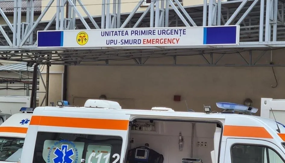 Imaginea prezintă o ambulanță parcată în fața unei clădiri, sub un panou pe care scrie "UNITATEA PRIMIRE URGENȚE UPU-SMURD EMERGENCY".