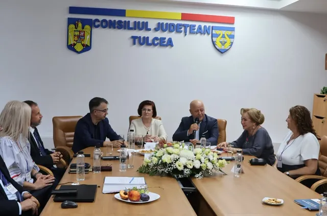 Șase persoane stau la o masă de conferințe sub un panou cu inscripția Consiliul Județean Tulcea, având un buchet de flori în centru.