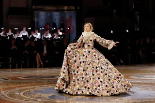 O femeie într-o rochie lungă, cu model floral și geometric, stă pe un podea decorată în fața unui grup de oameni așezați în umbră.