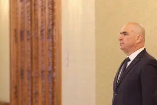 Un bărbat cu capul chel și costum închis la culoare stă în profil în fața unui perete și al unei uși de lemn.
