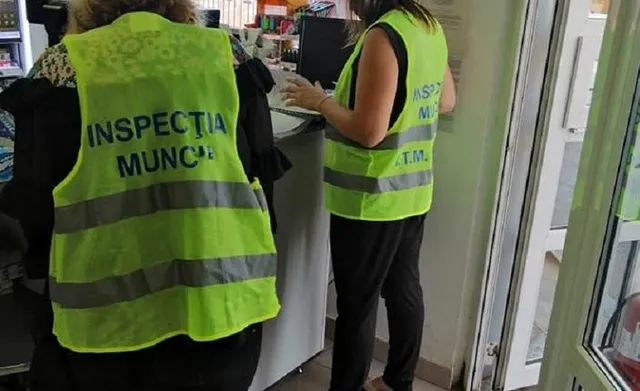 Două persoane purtând veste reflectorizante galben-verzui cu inscripții pe spate stau în fața unui birou cu un monitor de calculator.