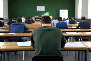 Imaginea prezintă o sală de clasă cu elevi așezați la bănci, văzuți din spate, cu un elev în prim-plan, având o foaie pe bancă.