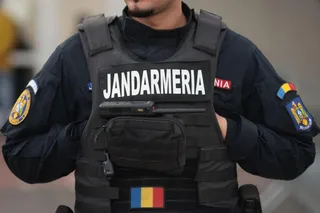 Un membru al jandarmiei poartă o vestă tactică neagră cu inscripția JANDARMERIA și un steag românesc pe partea inferioară.