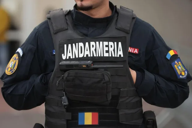 Un membru al jandarmiei poartă o vestă tactică neagră cu inscripția JANDARMERIA și un steag românesc pe partea inferioară.