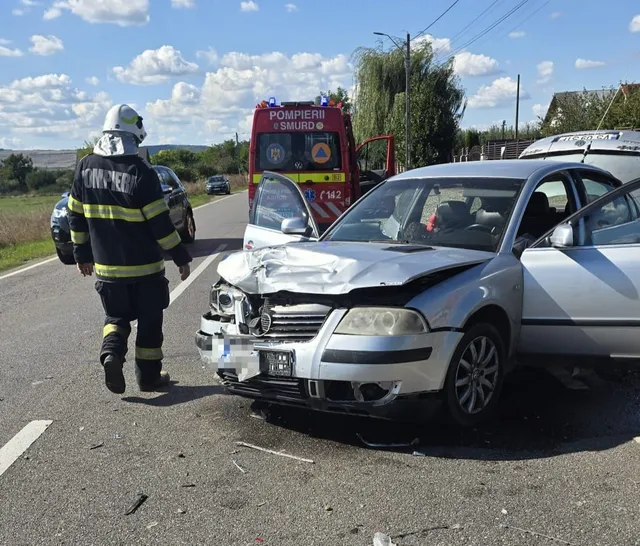 Un pompier în uniformă neagră cu benzi reflectorizante galbene stă lângă un autoturism argintiu cu partea frontală zdrobită pe un drum asfaltat.