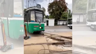 Un tramvai verde și alb circulă pe șine într-o zonă cu pavaj și vegetație sub un cer înnorat.