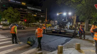 O echipă de lucrători în veste reflectorizante lucrează pe un drum asfaltat noaptea lângă un utilaj de pavaj și un drum de pietriș.