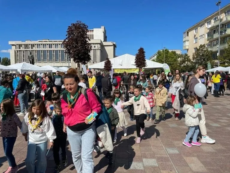 O mulțime traversează o piață cu corturi albe, postere expuse și clădiri în fundal, sub cer senin.