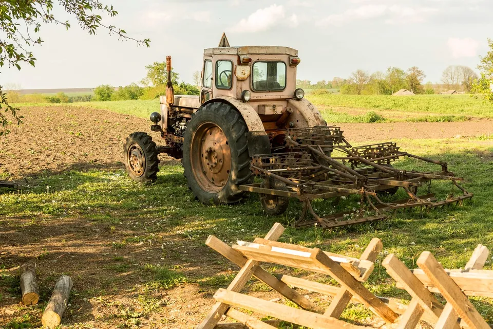 Un tractor de culoare deschisă cu un utilaj de arat este parcat pe un câmp lângă câteva bușteni și structuri din lemn.