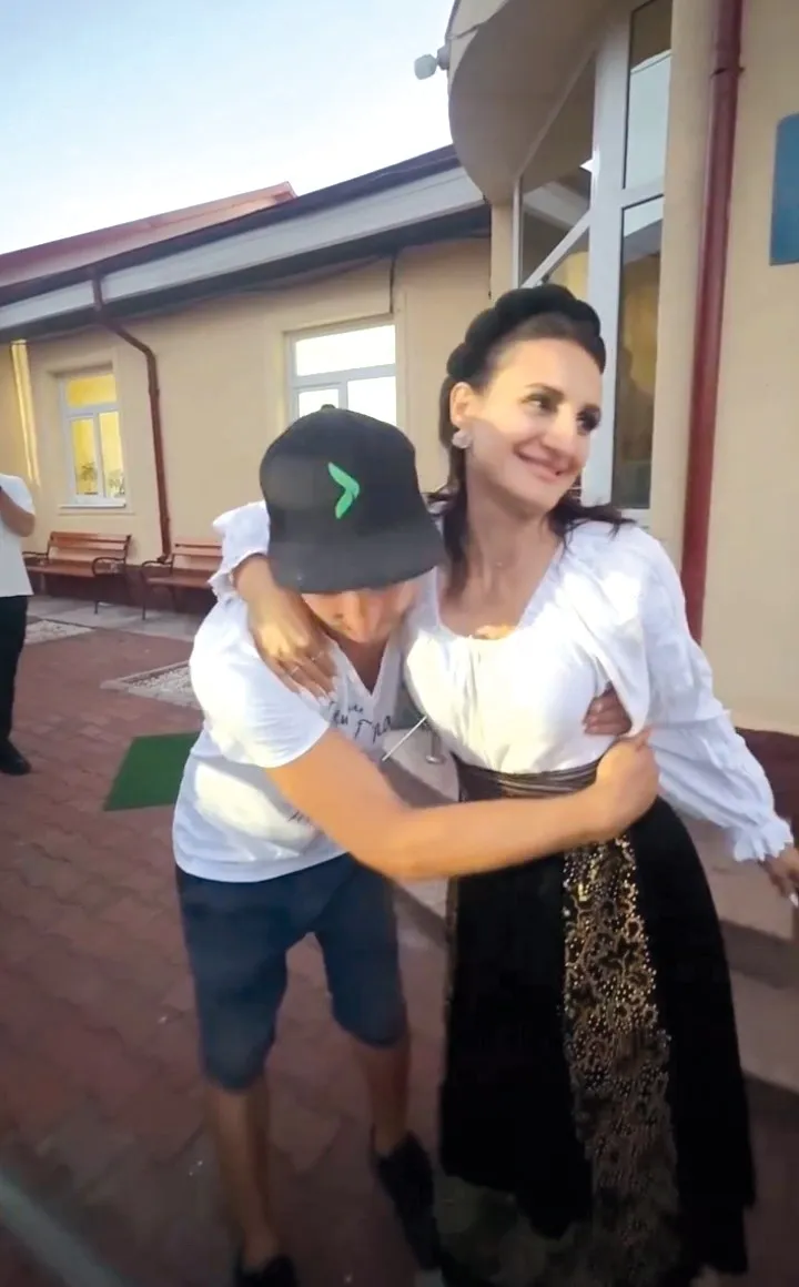 O femeie cu părul închis la culoare și o cămașă albă îmbrățișează un copil care poartă șapcă neagră și tricou alb.