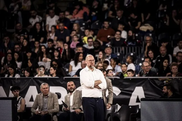 Un bărbat cu ochelari și cămașă albă stă cu brațele încrucișate în fața unui grup de oameni așezați pe tribunele unei săli.
