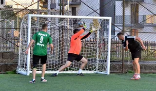 Un jucător în echipament portar de culoare portocalie sare spre o minge în fața unei porți de fotbal, în timp ce alți doi jucători îl urmăresc.
