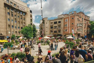 O mulțime de oameni stă la mese în aer liber într-o piață urbană înconjurată de clădiri înalte sub un cer cu nori.