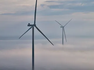 Două turbine eoliene înalte se ridică deasupra unui strat dens de ceață sub un cer înnorat.