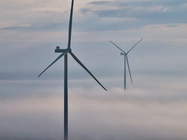 Două turbine eoliene înalte se ridică deasupra unui strat dens de ceață sub un cer înnorat.