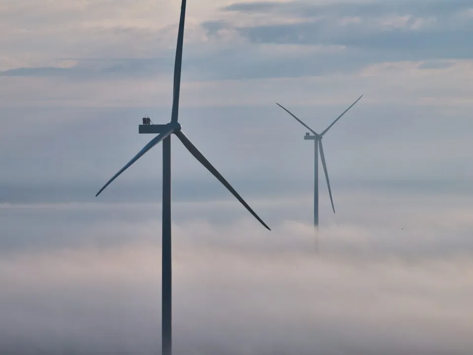 Două turbine eoliene înalte se ridică deasupra unui strat dens de ceață sub un cer înnorat.