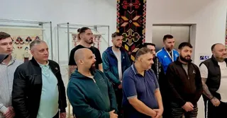Un grup de bărbați stă în picioare într-o încăpere cu vitrine de sticlă și un tapet decorativ pe perete.