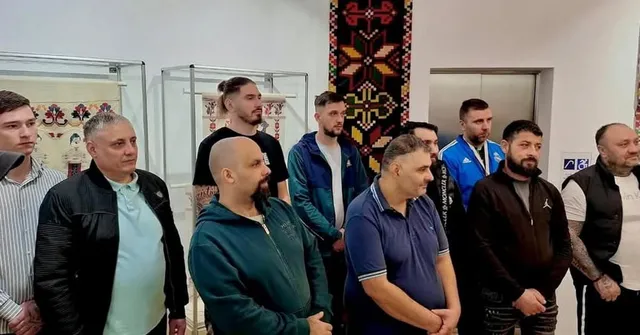 Un grup de bărbați stă în picioare într-o încăpere cu vitrine de sticlă și un tapet decorativ pe perete.
