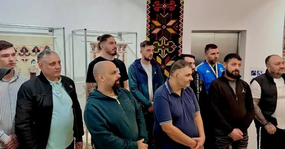 Un grup de bărbați stă în picioare într-o încăpere cu vitrine de sticlă și un tapet decorativ pe perete.