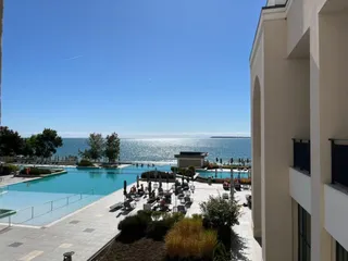 O piscină exterioară de formă neregulară este situată lângă o clădire albă, cu oameni așezați la mese și șezlonguri pe o terasă pavată.