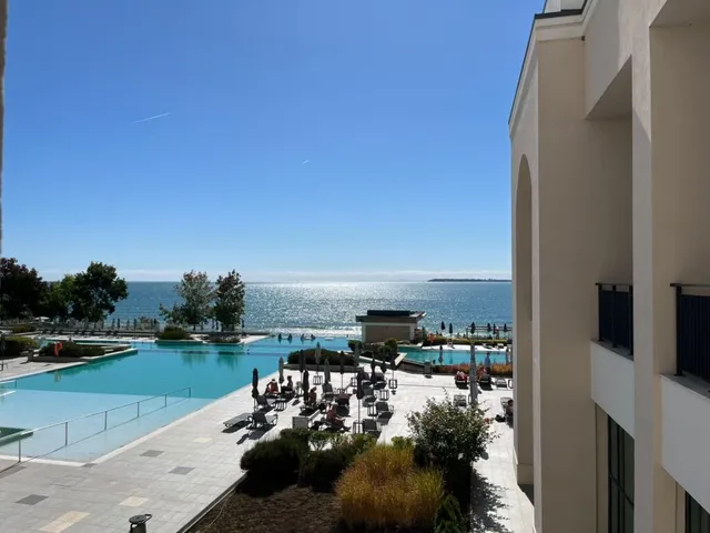 O piscină exterioară de formă neregulară este situată lângă o clădire albă, cu oameni așezați la mese și șezlonguri pe o terasă pavată.