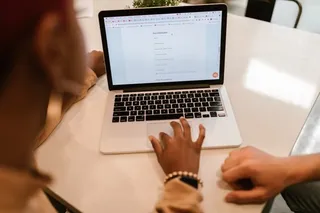 O persoană folosește un laptop deschis pe o masă albă, în timp ce o altă persoană stă lângă aceasta.