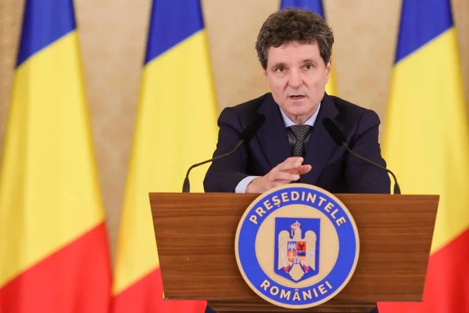 Un bărbat cu părul închis la culoare, purtând costum închis și cravată, stă în spatele unui podium maro cu un emblema circulară în față.