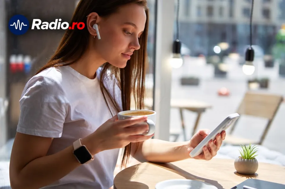 O femeie cu căști albe și ceas inteligent stă la o masă, ținând o cană și un telefon mobil într-o încăpere luminoasă.