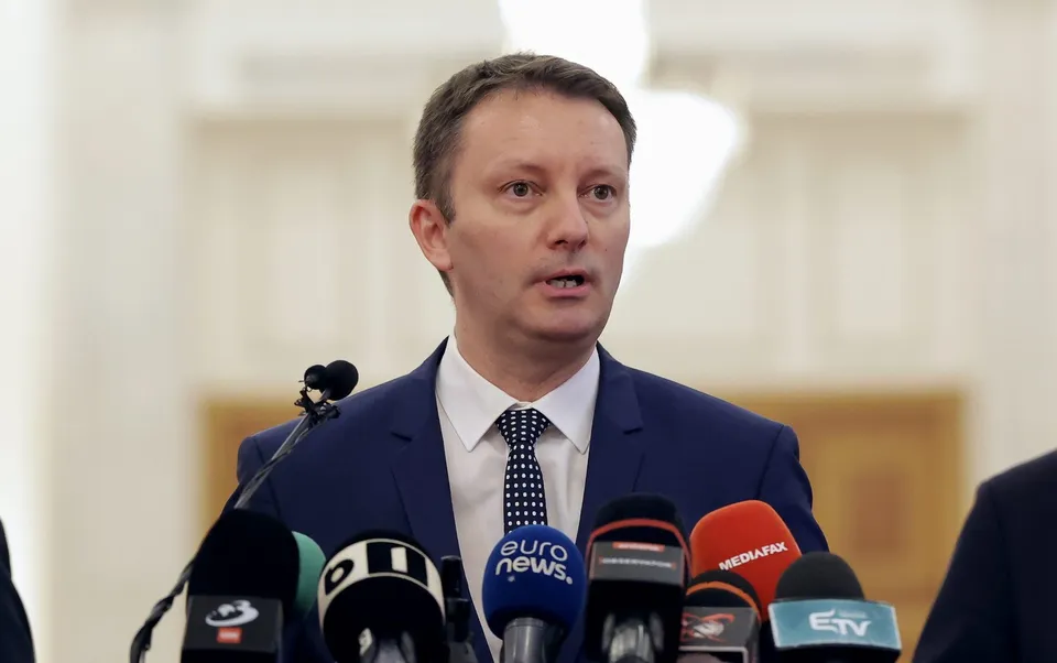 Un bărbat cu cămașă albă, cravată albastră cu puncte și sacou albastru vorbește în fața mai multor microfoane de presă.