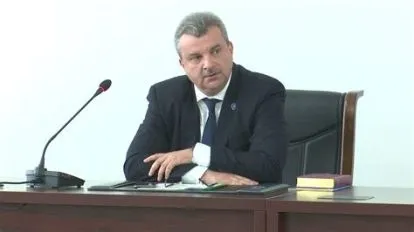 Un bărbat cu părul scurt și costum închis se așază la o masă de lemn lângă un microfon și un obiect dreptunghiular colorat.