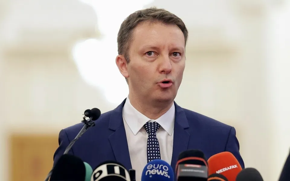 Un bărbat în costum albastru stă în fața mai multor microfoane, cu gura ușor deschisă.