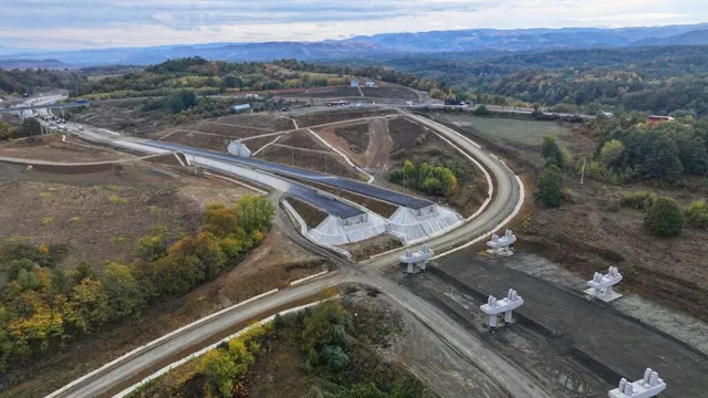 O vedere aeriană prezintă un șantier de construcții cu drumuri de acces, pământ excavat și structuri de beton albe pe un teren colinar.