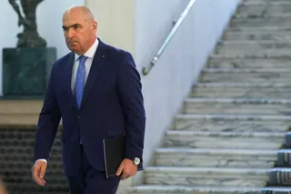 Un bărbat cu capul chel, purtând un costum albastru și cravată albastră, merge pe lângă o scară de marmură purtând un dosar negru.