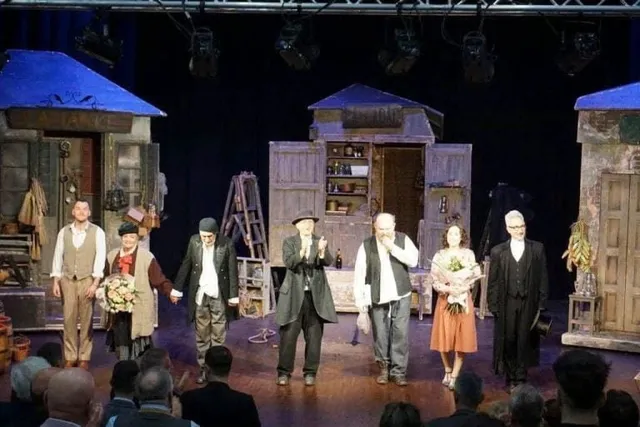 Oamenii stau pe o scenă de teatru în fața unui public, purtând diverse costume, lângă decoruri care reprezintă structuri de lemn.